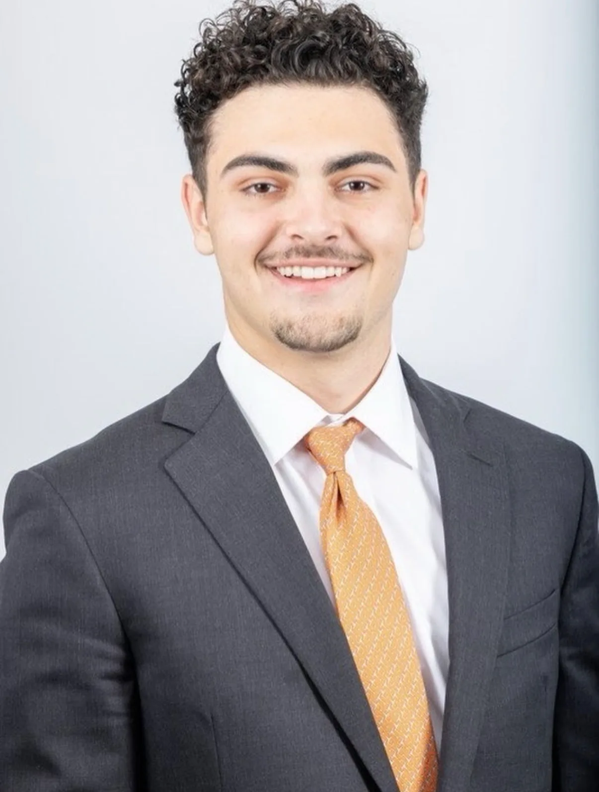 Professional headshot of Ian Massaro wearing a dark suit, white shirt, and orange tie against a light background.