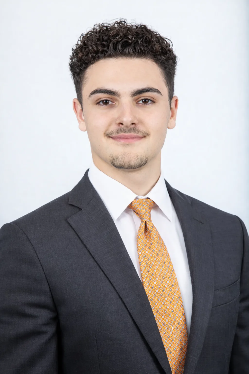 Professional headshot of Ian Massaro wearing a dark suit, white shirt, and orange tie against a light background.